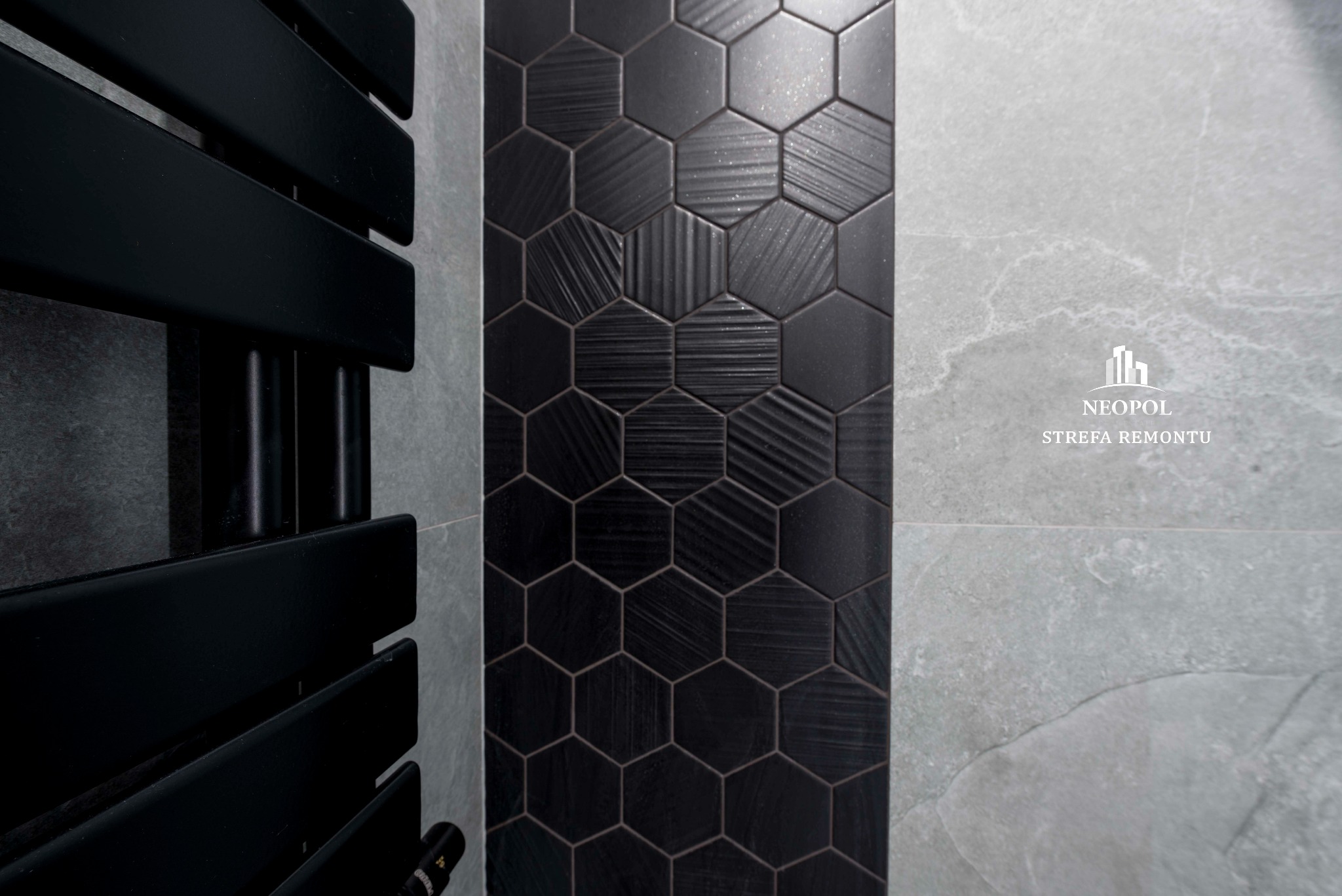 Fragment łazienki z czarnym grzejnikiem drabinkowym i ścianą wyłożoną heksagonalnymi, czarnymi płytkami z różnymi fakturami, sąsiadującą z szarą płytą imitującą kamień. Widoczne logo 'NEOPOL STREFA...
