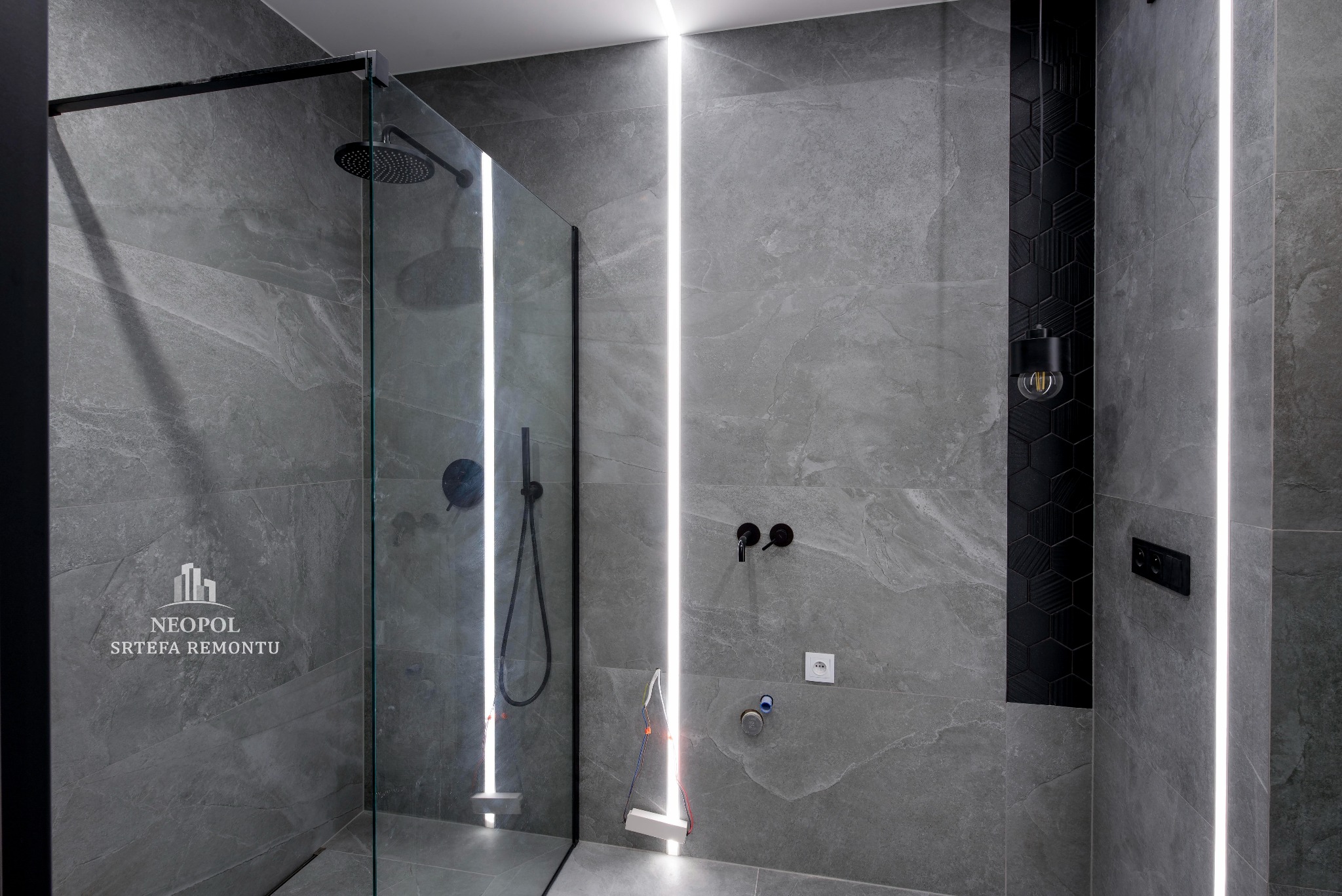 Nowoczesna łazienka z szarymi płytkami imitującymi kamień, kabina prysznicowa typu walk-in z czarnymi elementami, pionowe oświetlenie LED w ścianie, dekoracyjny panel z czarnych heksagonów...