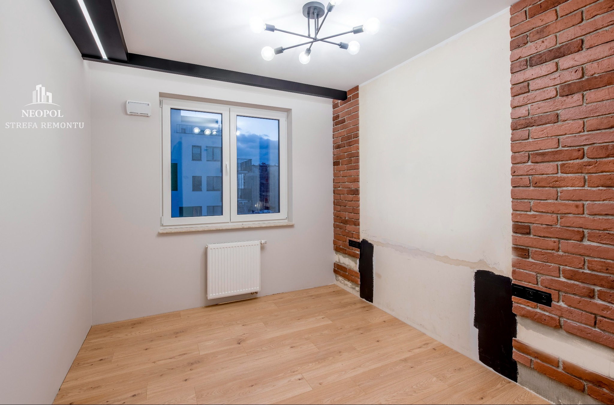 Pokój w trakcie aranżacji w Warszawie: ściana z cegły dekoracyjnej, fragment ściany przygotowany pod malowanie z widocznym gruntem, drewniana podłoga, okno z widokiem na budynek, nowoczesna lampa...