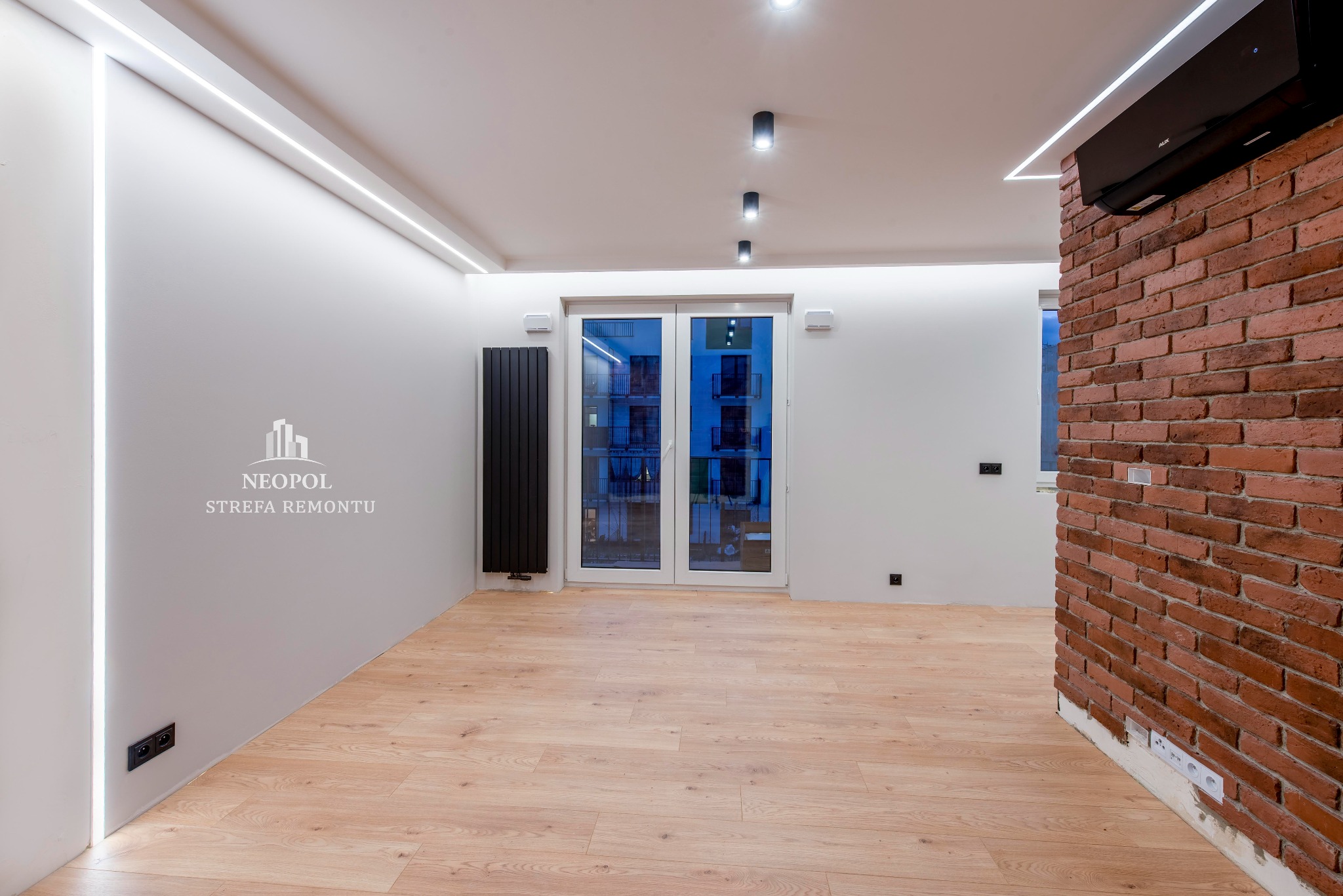 Wnętrze po remoncie z ceglaną ścianą, oświetleniem LED w suficie i przy ścianach, nowoczesnym grzejnikiem i klimatyzatorem, widok na okno balkonowe.