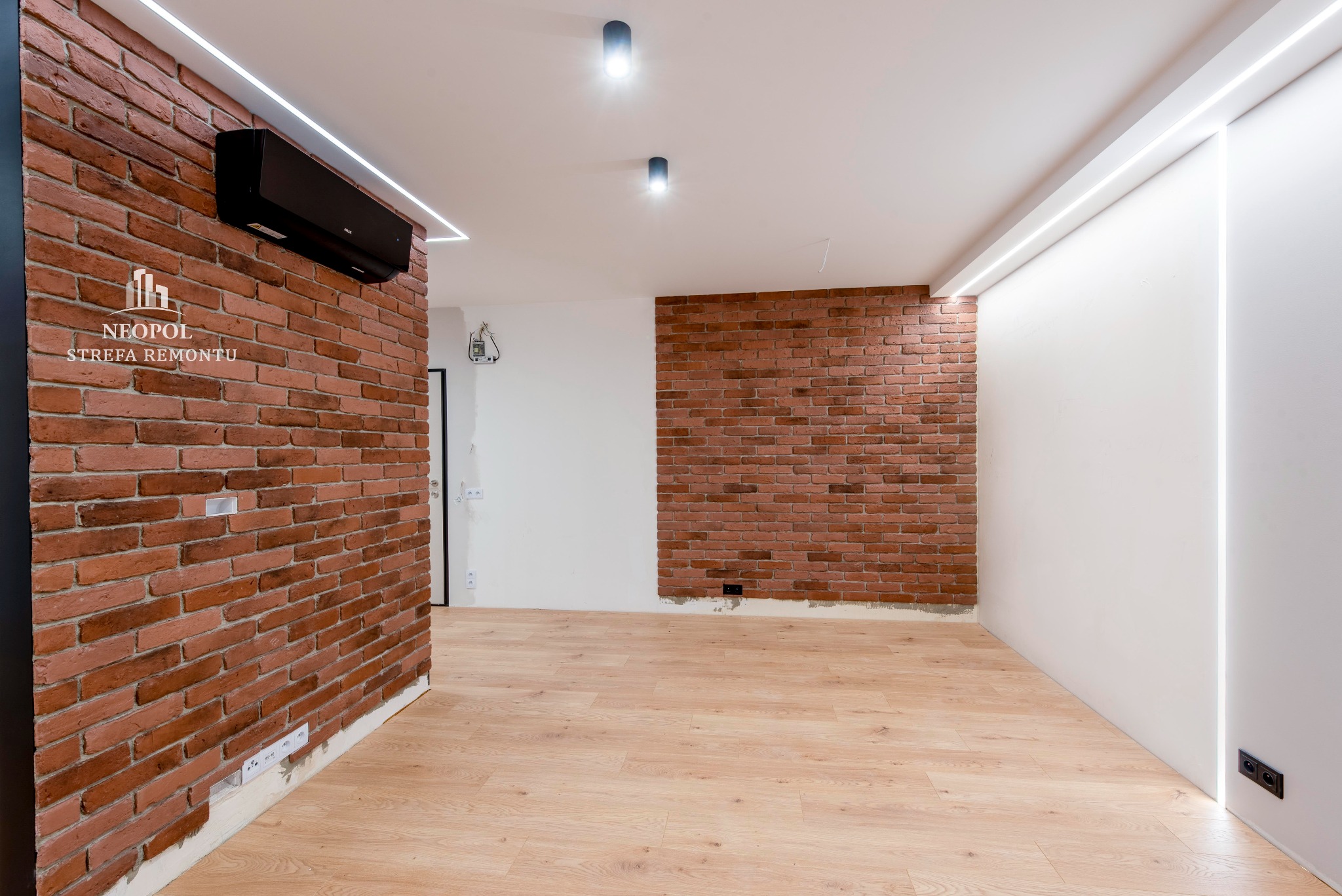 Wykończone wnętrze z ceglaną ścianą, klimatyzacją i oświetleniem LED, widoczny fragment logo firmy remontowej na ścianie.