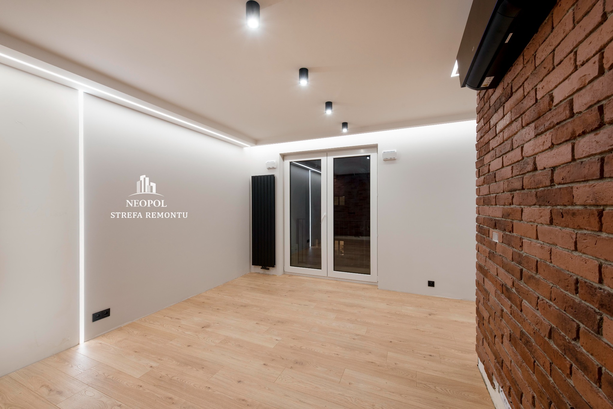Puste pomieszczenie po remoncie z panelami podłogowymi, ceglaną ścianą, grzejnikiem żeberkowym, oświetleniem sufitowym, klimatyzacją i logo firmy remontowej na ścianie.