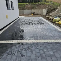 Ułożona, częściowo mokra, szara kostka brukowa z białym obramowaniem przy białym budynku. W tle skarpa z zielenią i betonowym murem oporowym.