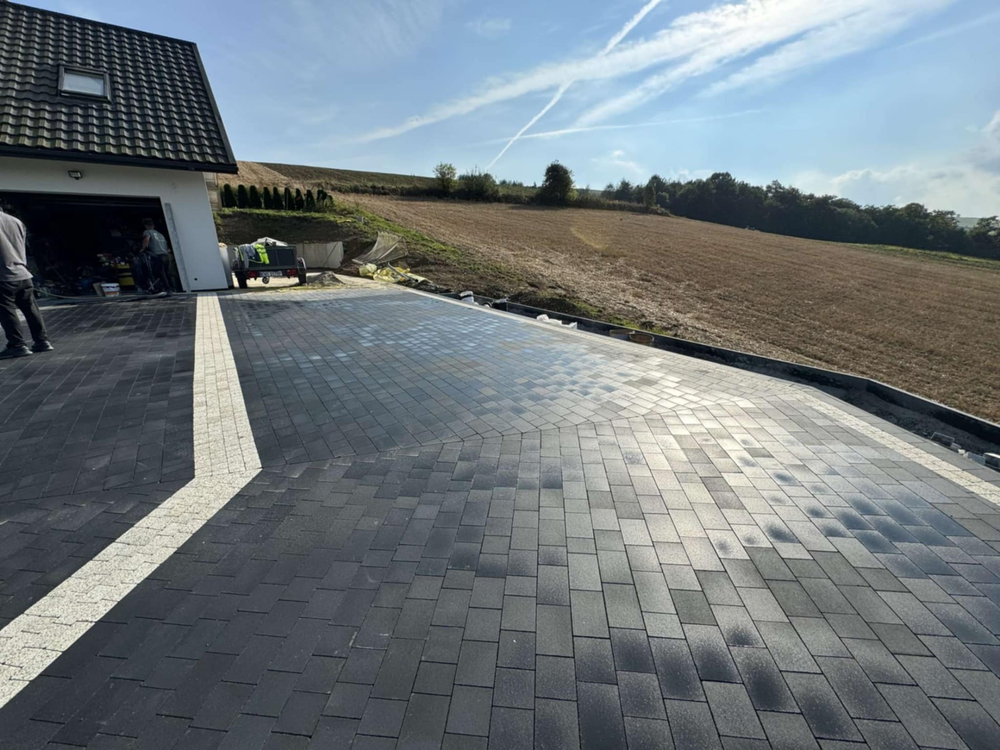 Ułożona, grafitowa kostka brukowa na podjeździe, z jasnym pasem oddzielającym od domu. W tle widoczny krajobraz pola i błękitne niebo z chmurami.
