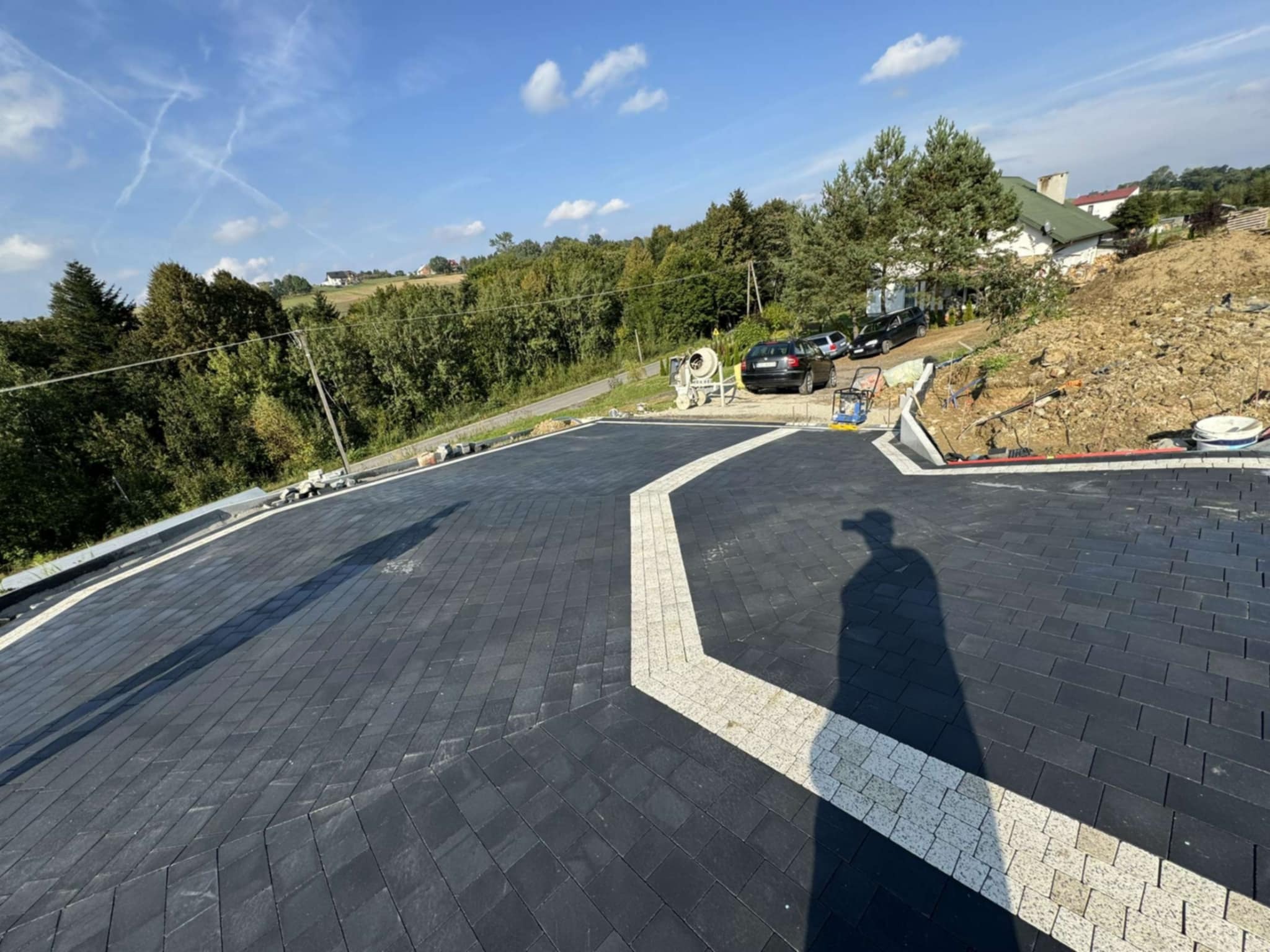 Ułożona, ciemnoszara kostka brukowa z jasnym pasem w kształcie litery L na podjeździe. W tle zieleń, dom i cień osoby.