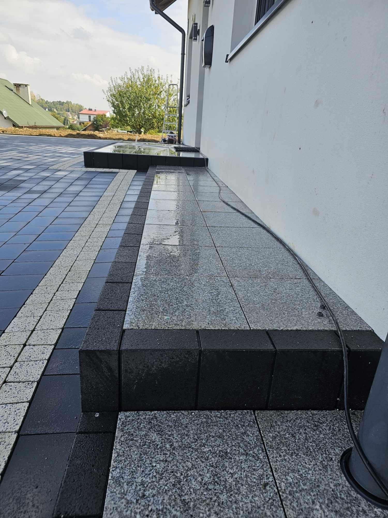 Ułożona kostka brukowa w odcieniach szarości i czerni, prowadząca do ściany budynku. Widoczne schody z kostki i mokra nawierzchnia.