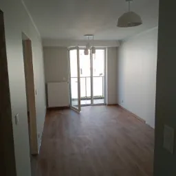 Pusta, jasna przestrzeń po remoncie z panelami podłogowymi, białymi ścianami, grzejnikiem i otwartymi drzwiami balkonowymi, widoczne dwie lampy sufitowe.