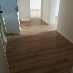 Wnętrze mieszkania z nową, drewnopodobną podłogą laminowaną, widoczne drzwi wewnętrzne i białe ściany w stanie surowym.