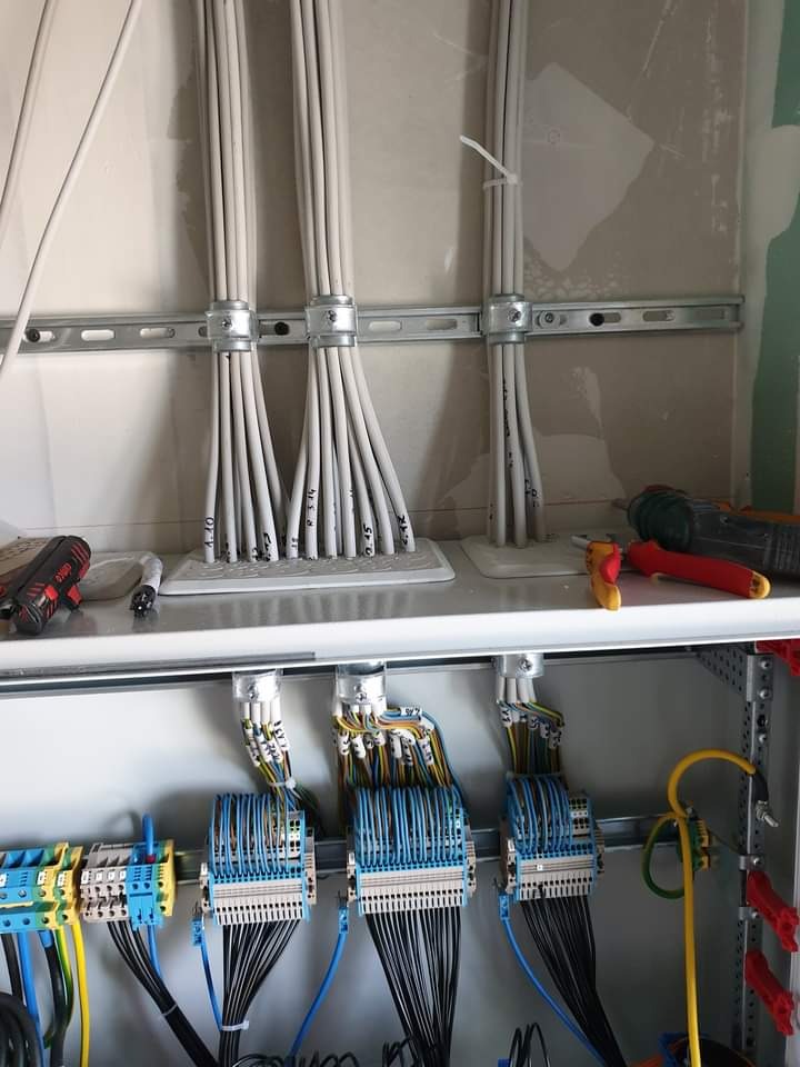 Szczegółowe ujęcie rozdzielnicy elektrycznej z licznymi przewodami oznaczonymi numerami, uporządkowanymi w wiązki i podłączonymi do listew zaciskowych, widoczne narzędzia pracy na górnej półce.