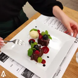 Elegancko podany deser: dwa kawałki brownie polane sosem owocowym, udekorowane świeżymi malinami, bor&oacute;wkami, zielonymi winogronami i miętą, obok gałka lod&oacute;w waniliowych na kwadratowym talerzu,...