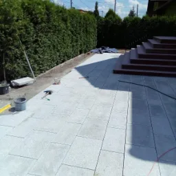 Jasnoszary taras z kamiennych płyt w trakcie budowy, widoczne narzędzia, wiaderka z zaprawą, poziomica, w tle zielony żywopłot i schody wejściowe do domu.