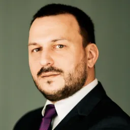 Portret mężczyzny w ciemnym garniturze i fioletowym krawacie, patrzącego wprost, na neutralnym tle.