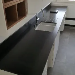 Długa, czarna, matowa powierzchnia blatu kuchennego z białym zlewem i baterią, widok na miasto przez okno w tle.