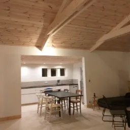 Wnętrze domu z drewnianym sufitem podwieszanym z widocznymi belkami stropowymi, otwarta kuchnia z białymi szafkami, stół z krzesłami.