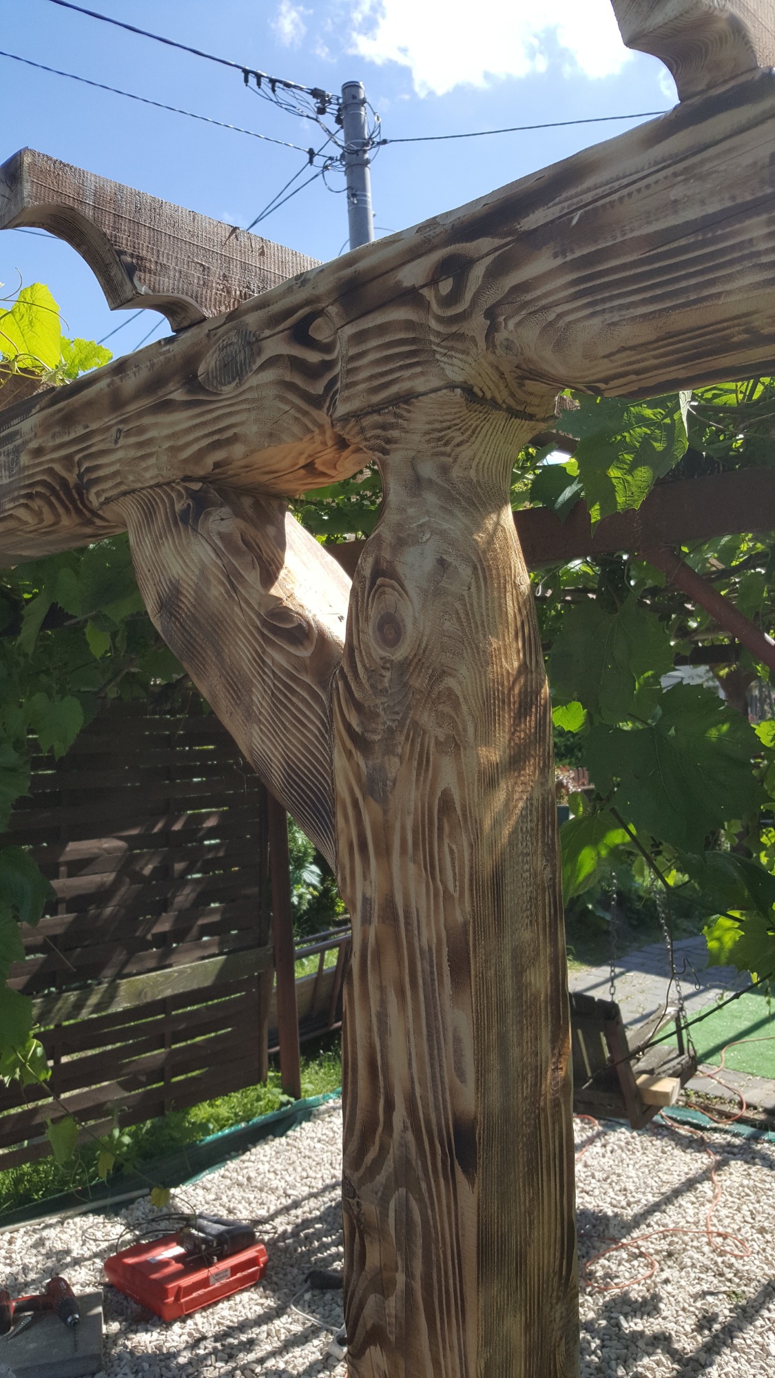 Fragment drewnianej pergoli z wyraźnym rysunkiem słojów, widoczne połączenia belek, w tle zieleń i narzędzia na żwirowym podłożu.