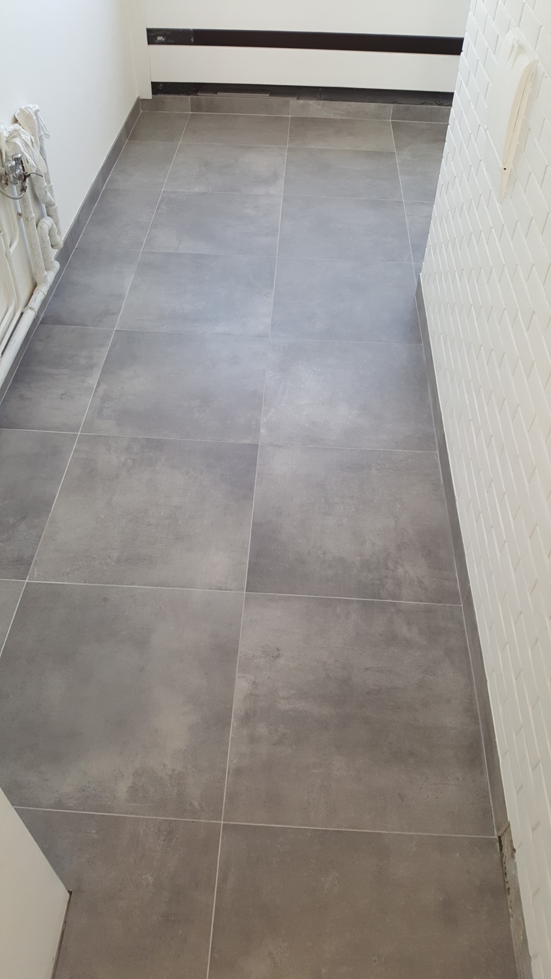 Ułożona, szara, matowa podłoga z płytek ceramicznych w korytarzu, widok z góry, biała ściana z cegiełek po prawej stronie.