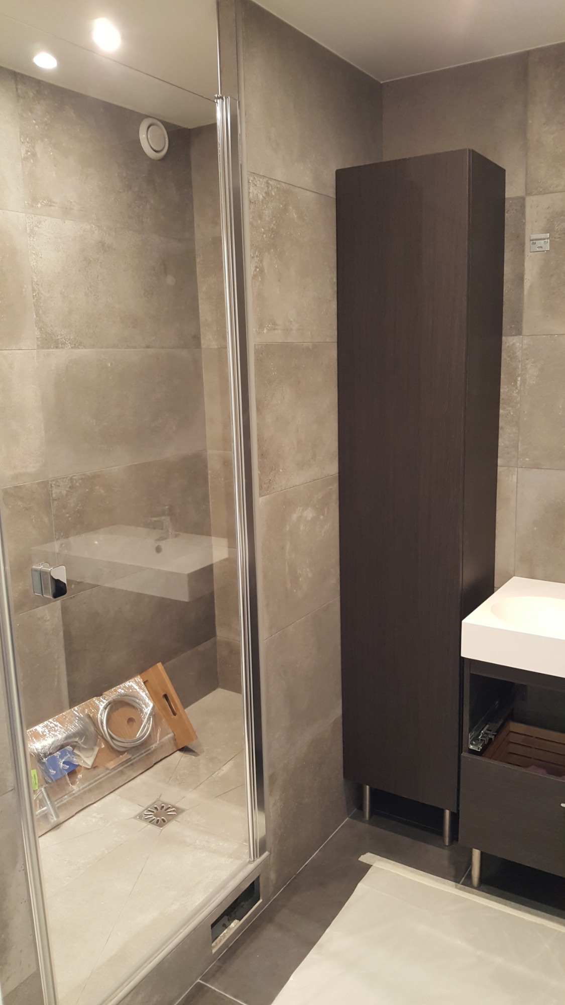 Nowoczesna łazienka w trakcie wykańczania: kabina prysznicowa z transparentnymi drzwiami, podwieszana umywalka, wysoka szafka z ciemnego drewna, narzędzia i materiały budowlane na podłodze.