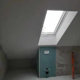 Poddasze w trakcie remontu z oknem dachowym, zabudową z płyty gipsowo-kartonowej z instalacją sanitarną i poziomicą opartą o ścianę.