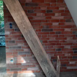 Ściana z cegły klinkierowej w odcieniach czerwieni i brązu, częściowo zasłonięta przez drewnianą belkę pod kątem, widoczne gniazdka elektryczne i fragment podłogi z płytek imitujących drewno.