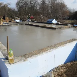 Wylany fundament pod dom z izolacją termiczną z białego styropianu, widoczna butelka z napojem i pracownicy w tle.