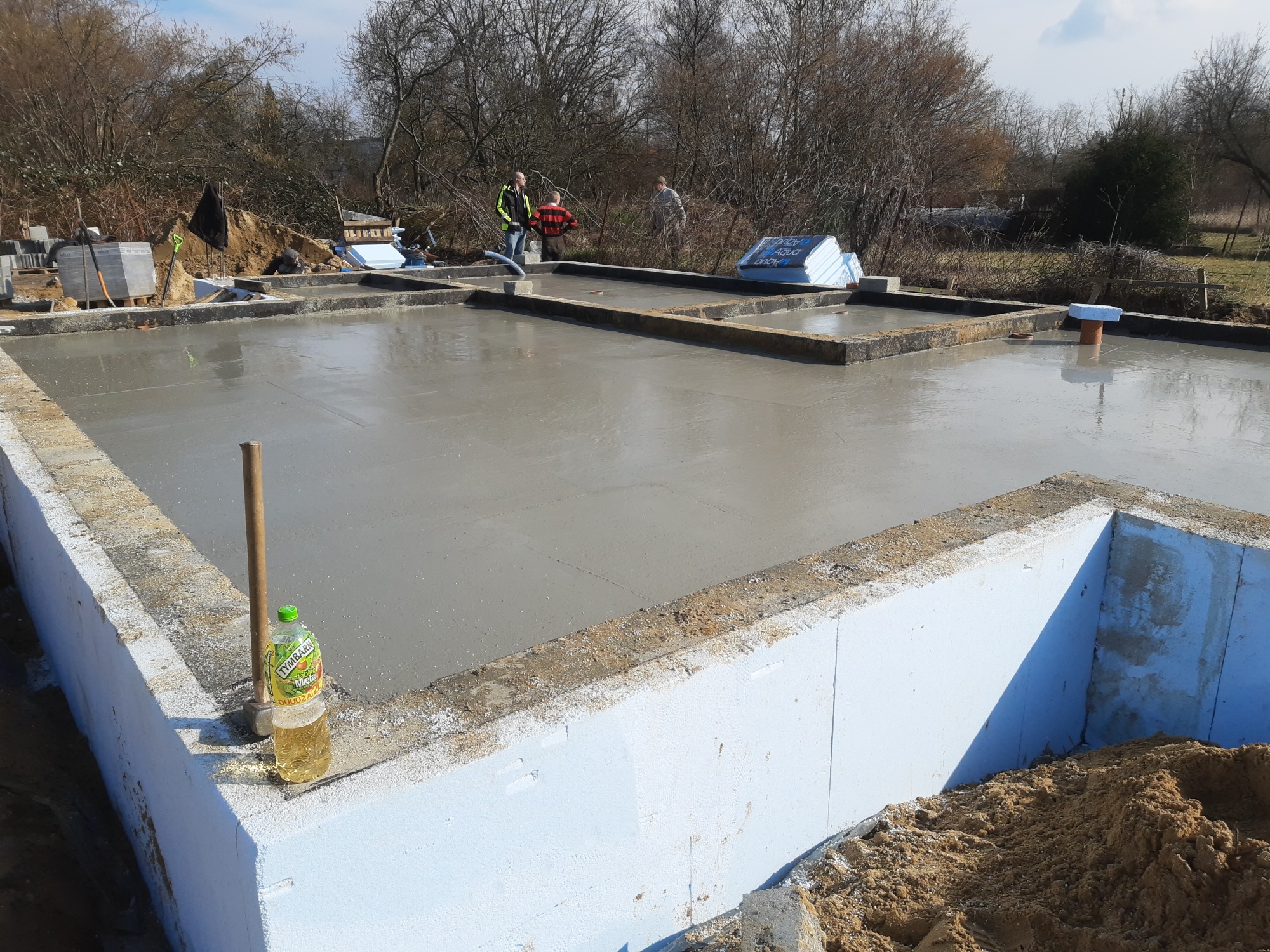 Wylany fundament pod dom z izolacją termiczną z białego styropianu, widoczna butelka z napojem i pracownicy w tle.