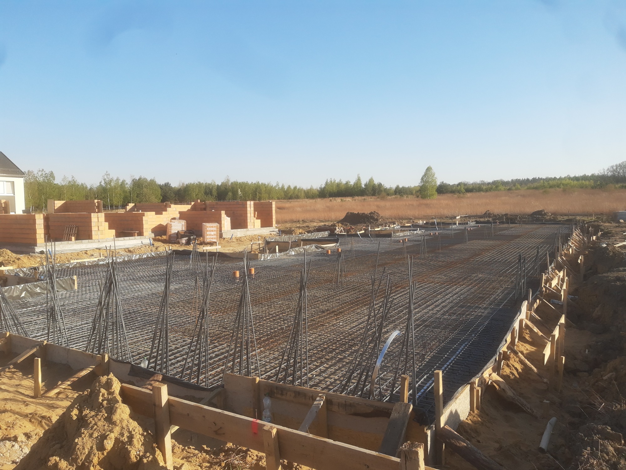 Zbrojenie fundamentu domu jednorodzinnego z widocznymi elementami szalunku i częściowo wymurowanymi ścianami z czerwonej cegły w tle.