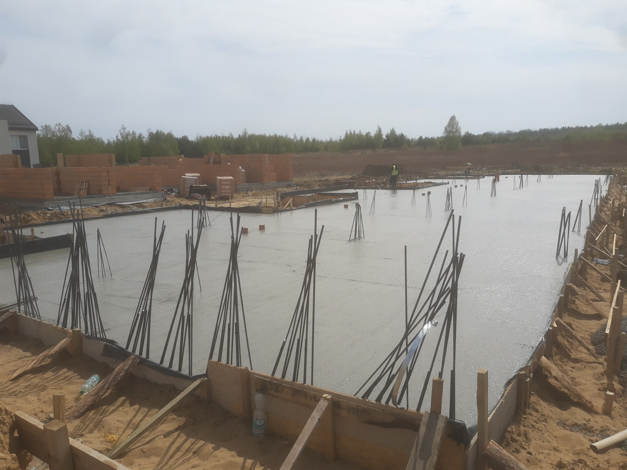 Zalany betonowy fundament z wystającymi prętami zbrojeniowymi, drewnianym szalunkiem i stosem bloczków silikatowych w tle.