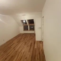 Poddasze po remoncie z drewnianą podłogą, białymi ścianami i dwoma oknami dachowymi, widoczna instalacja elektryczna sufitowa.