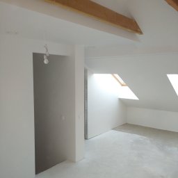 USŁUGI OGÓLNOBUDOWLANE Marek Ratajczak - Wnętrze poddasza po szpachlowaniu i malowaniu na biało, widoczne drewniane belki stropowe, okna dachowe i surowa podłoga, żarówka na kablu zwisająca z sufitu w przejściu.