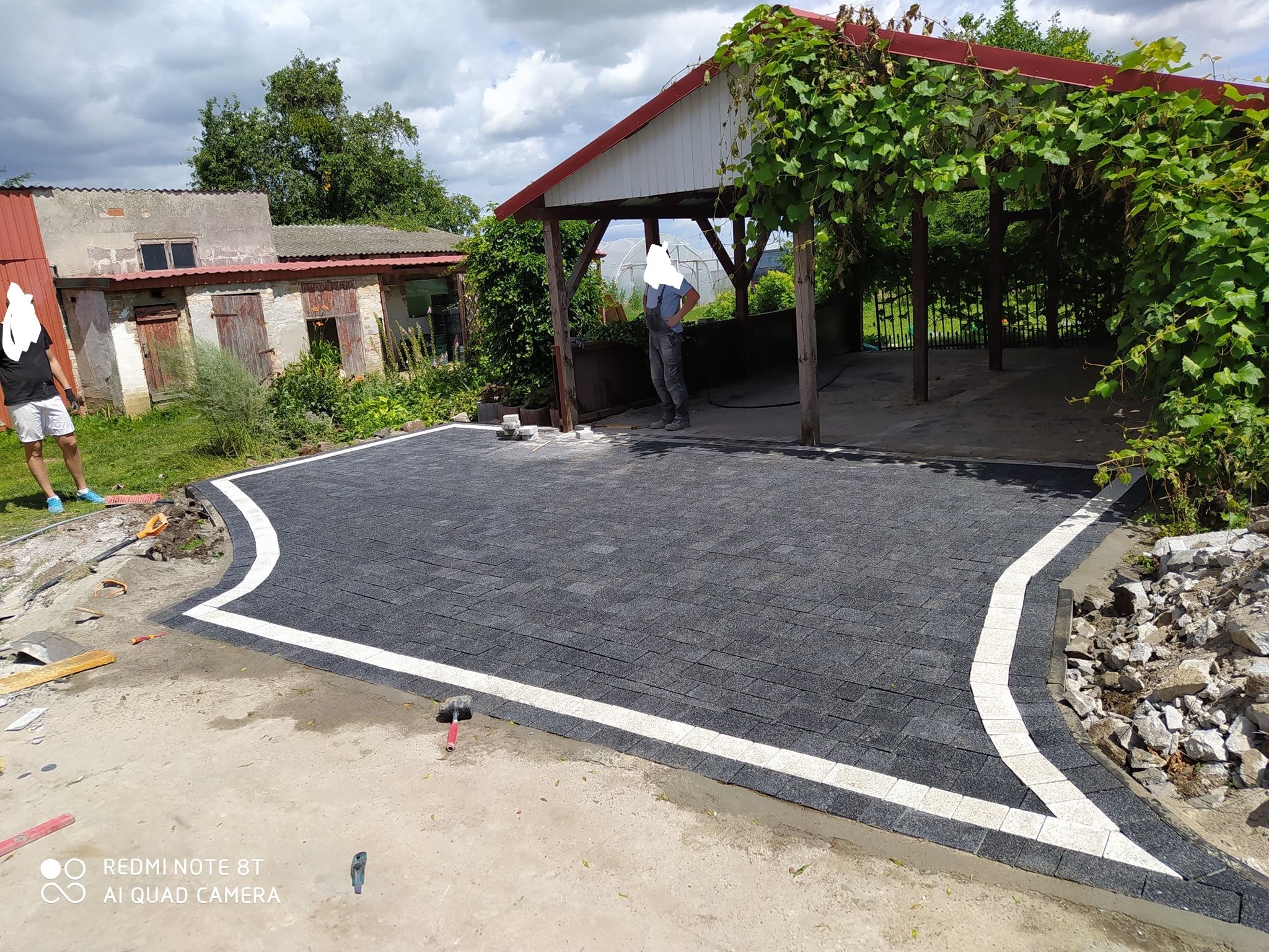 Częściowo ułożona szara kostka brukowa z białym obramowaniem na posesji, widoczne narzędzia i pracownicy, w tle budynek gospodarczy i altana porośnięta winoroślą.