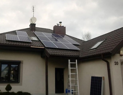 Montaż paneli słonecznych na brązowym dachu domu jednorodzinnego, widoczna drabina oparta o ścianę, częściowo zamontowane panele i jeden panel oparty o ścianę domu.