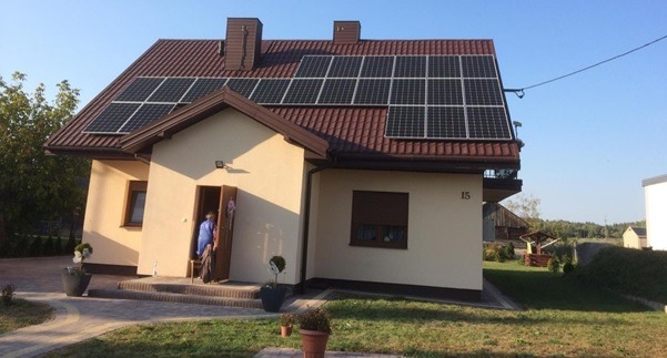 Dom jednorodzinny z brązowym dachem i panelami słonecznymi, numer domu 15, osoba w drzwiach, zadbany trawnik i dekoracyjne rośliny w donicach.