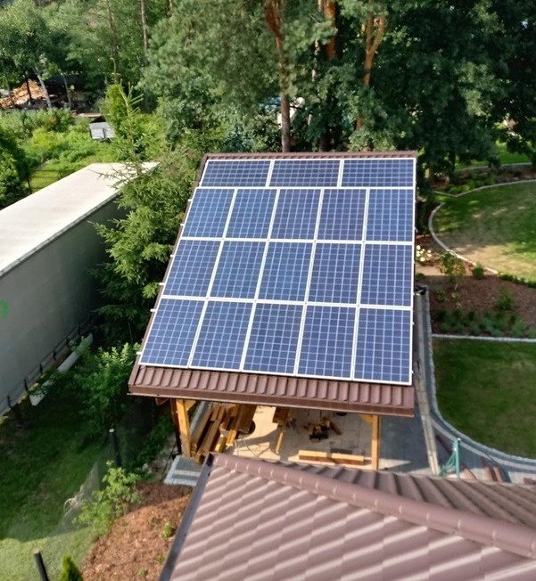 Instalacja paneli słonecznych na drewnianej konstrukcji w ogrodzie w Białymstoku, widok z góry na brązowy dach sąsiedniego budynku i zieleń drzew.