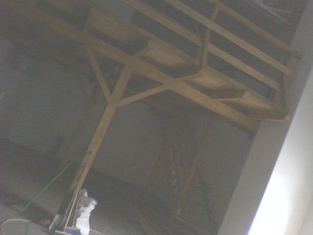 Konstrukcja drewniana poddasza z widocznymi belkami i elementami poszycia w trakcie docieplenia, widok z dołu w słabym oświetleniu.