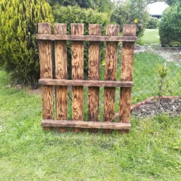 Ręcznie wykonany, opalany drewniany płotek ogrodowy na trawniku, z siatką ogrodzeniową w tle.