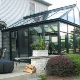 Szklana oranżeria z czarną ramą, przylegająca do domu, z widocznym tarasem z meblami ogrodowymi i ozdobnymi krzewami.