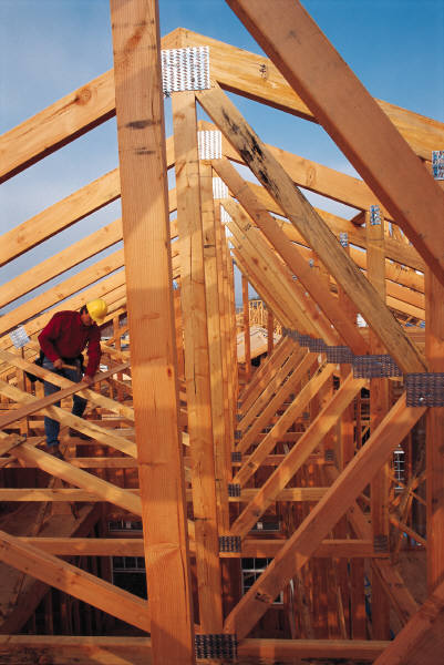 Konstrukcja drewniana dachu w trakcie budowy, widoczne elementy mocujące i pracownik w kasku, jasne drewno i niebieskie niebo w tle.