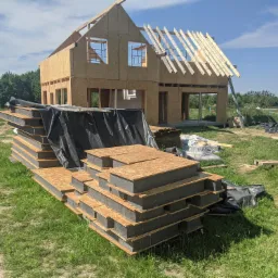 Wznoszenie dwukondygnacyjnego budynku z drewnianą konstrukcją szkieletową; widoczne przygotowane panele warstwowe z izolacją termiczną na pierwszym planie.