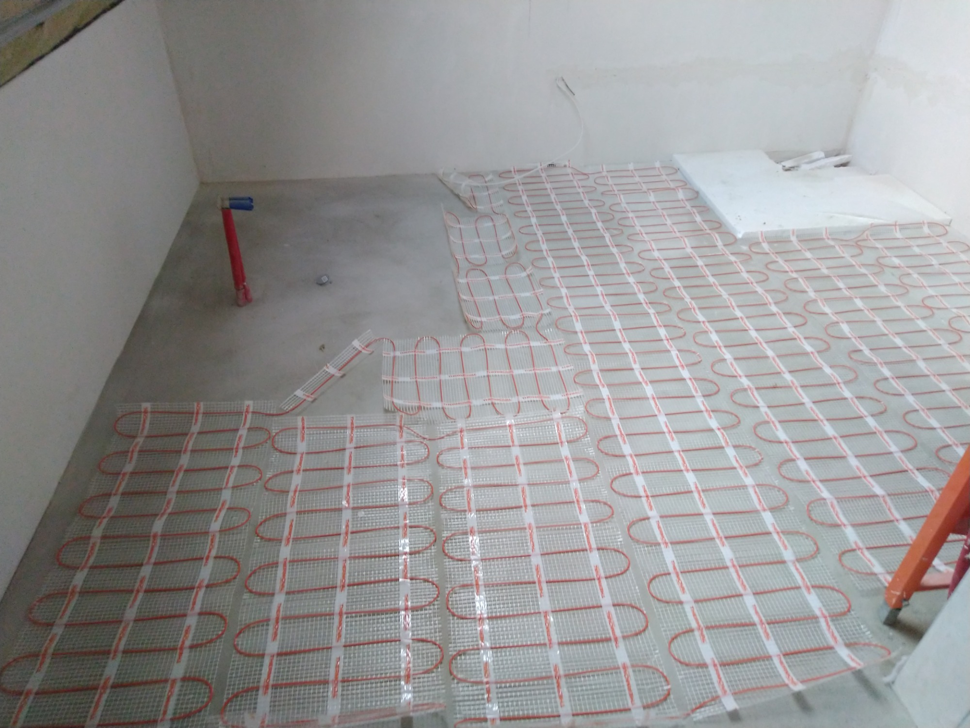 Rozłożona mata grzewcza z czerwonymi przewodami na szarej posadzce w trakcie montażu, widoczne fragmenty niepokrytej podłogi i rury przy ścianie.
