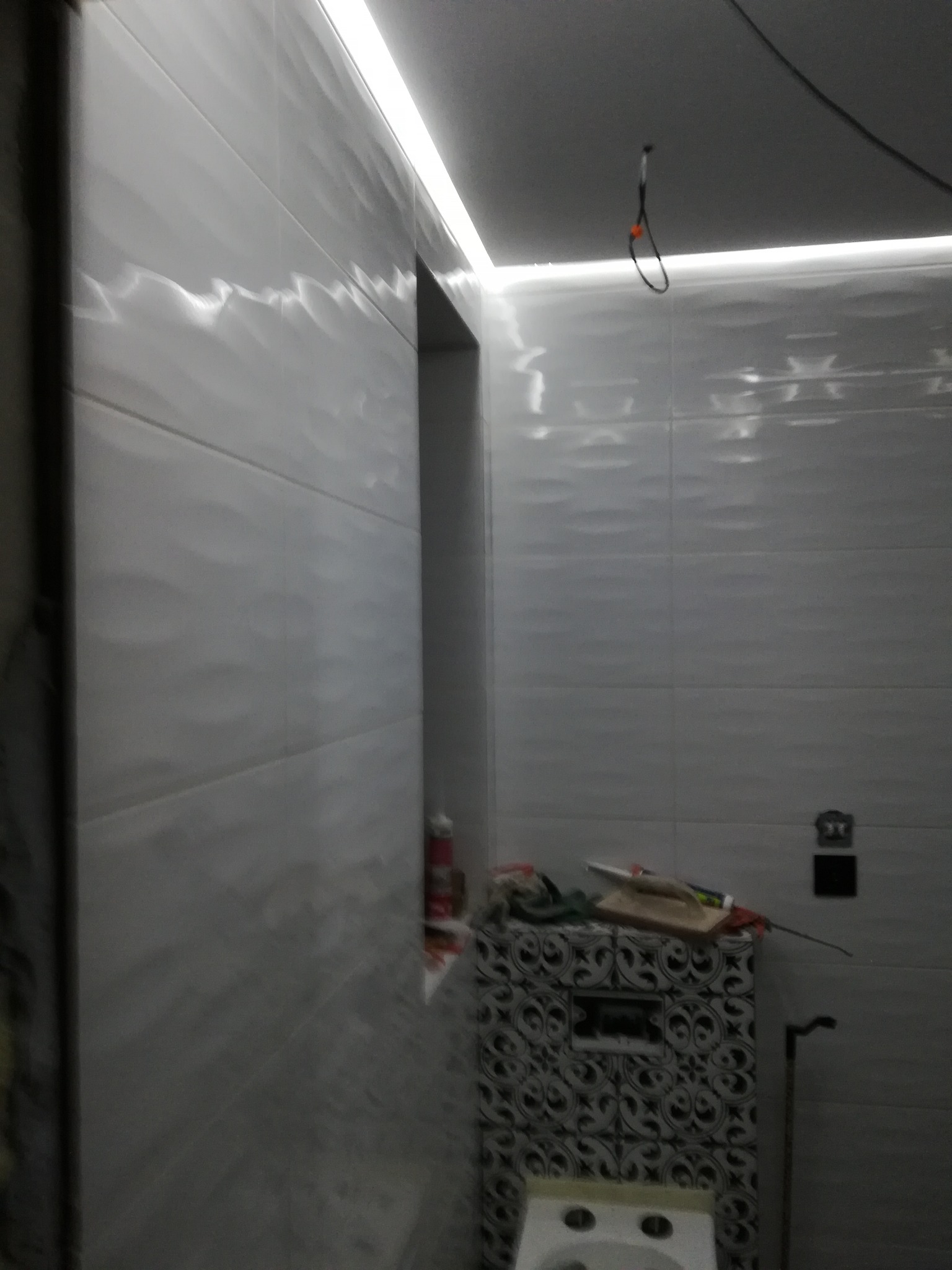 Ściana w łazience wyłożona białymi, błyszczącymi płytkami z delikatnym wzorem fal, oświetlona górnym światłem LED. Widoczny fragment obudowy stelaża WC z czarno-białym wzorem oraz narzędzia...