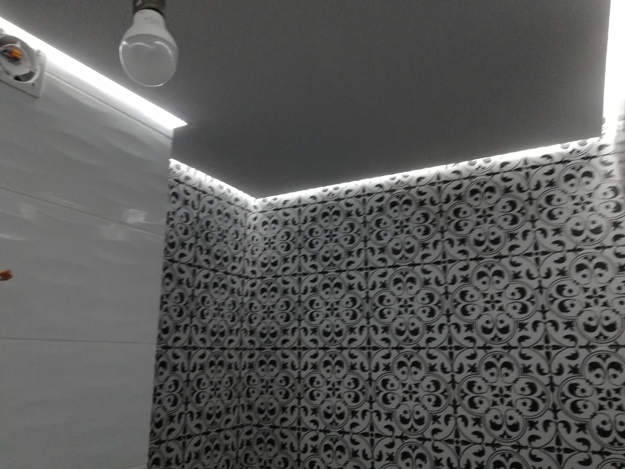 Białe płytki ścienne z dekoracyjnym wzorem, oświetlone listwą LED pod sufitem, widok na narożnik pomieszczenia z żarówką.