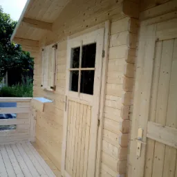 Drewniana altana z tarasem, jasne drewno, widoczne detale konstrukcyjne, okiennice i skrzynka na kwiaty.