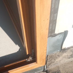 Grzegorz Pepliński - Fragment okna z drewnianą ramą, uszczelnionego taśmą izolacyjną i obróbką blacharską, zamontowanego w ścianie. Widoczne detale montażu i materiały.