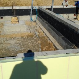 Grzegorz Pepliński - Fundamenty domu w trakcie budowy: izolacja pionowa, styropian, wystające rury. W tle hałda ziemi i częściowo widoczny budynek w stanie surowym otwartym.