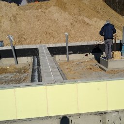 Grzegorz Pepliński - Fundamenty domu w trakcie budowy z widocznym ociepleniem, izolacją przeciwwilgociową i pracującym robotnikiem. W tle hałda piachu i żuraw budowlany.