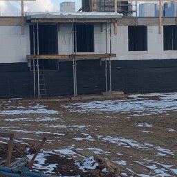 Grzegorz Pepliński - Budowa domu w trakcie realizacji: ściany z białych bloczków, czarna izolacja, rusztowania, podpory. Na pierwszym planie stos niebieskich podpór budowlanych.