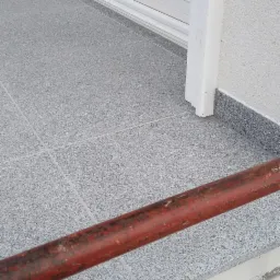 Szary, kwadratowy gres na balkonie z widoczną, rdzawą barierką i białymi drzwiami balkonowymi.