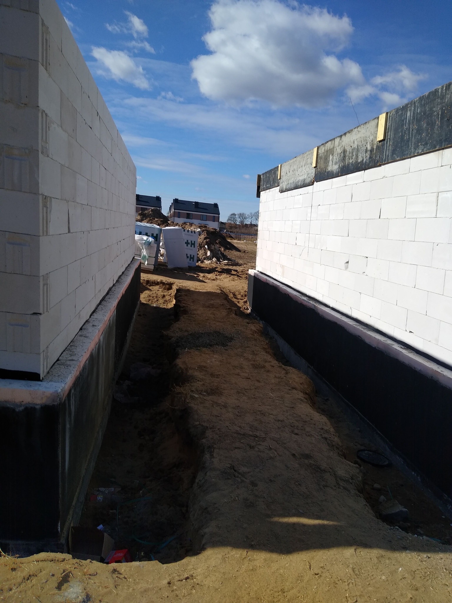 Budowa domu: wzniesione ściany z białych bloczków betonowych, zabezpieczone czarną izolacją przeciwwilgociową, widoczny wykop fundamentowy i materiały budowlane w tle.