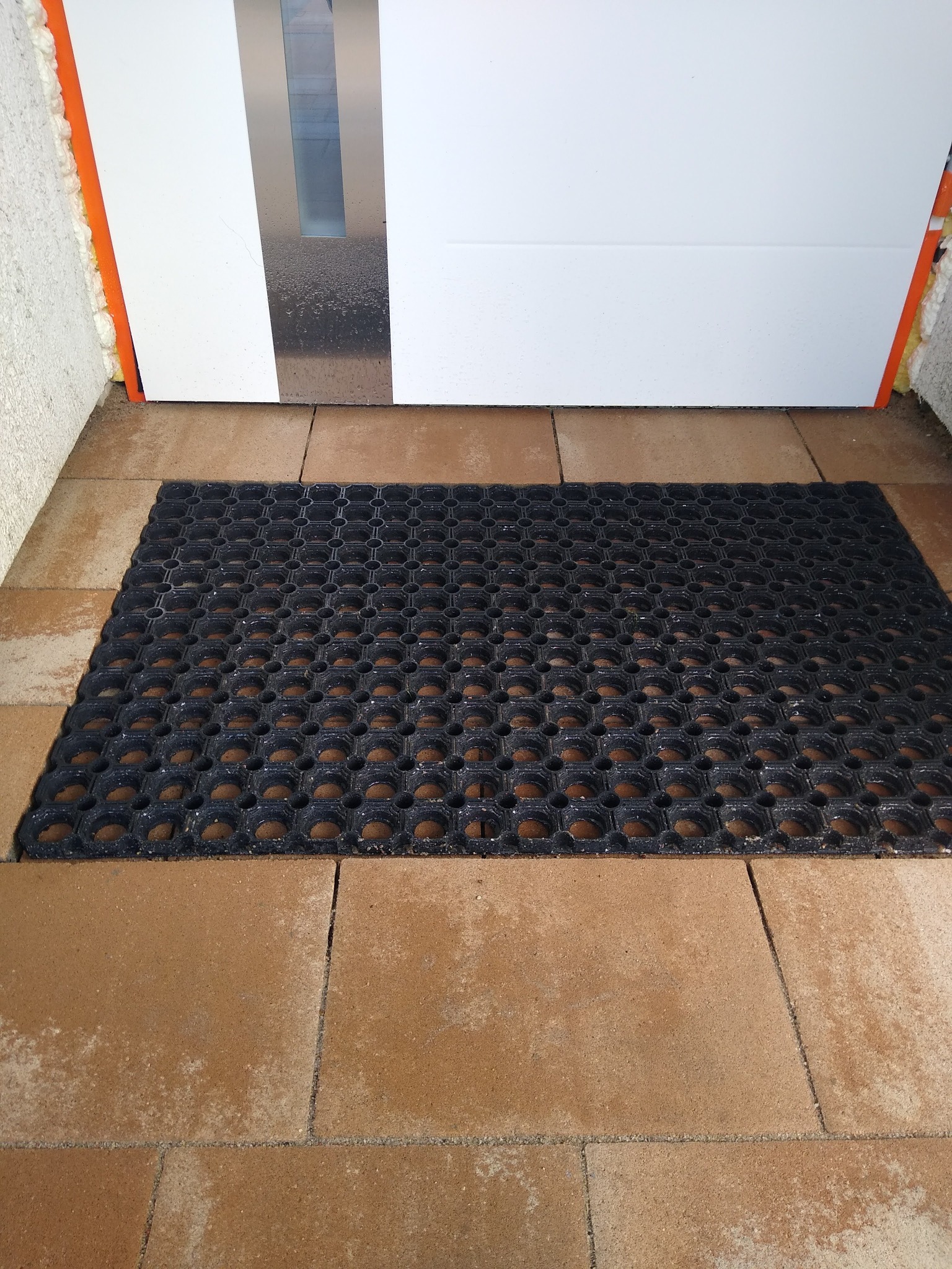 Brązowa kostka brukowa przed białymi drzwiami wejściowymi z czarną, gumową wycieraczką z okrągłymi otworami.