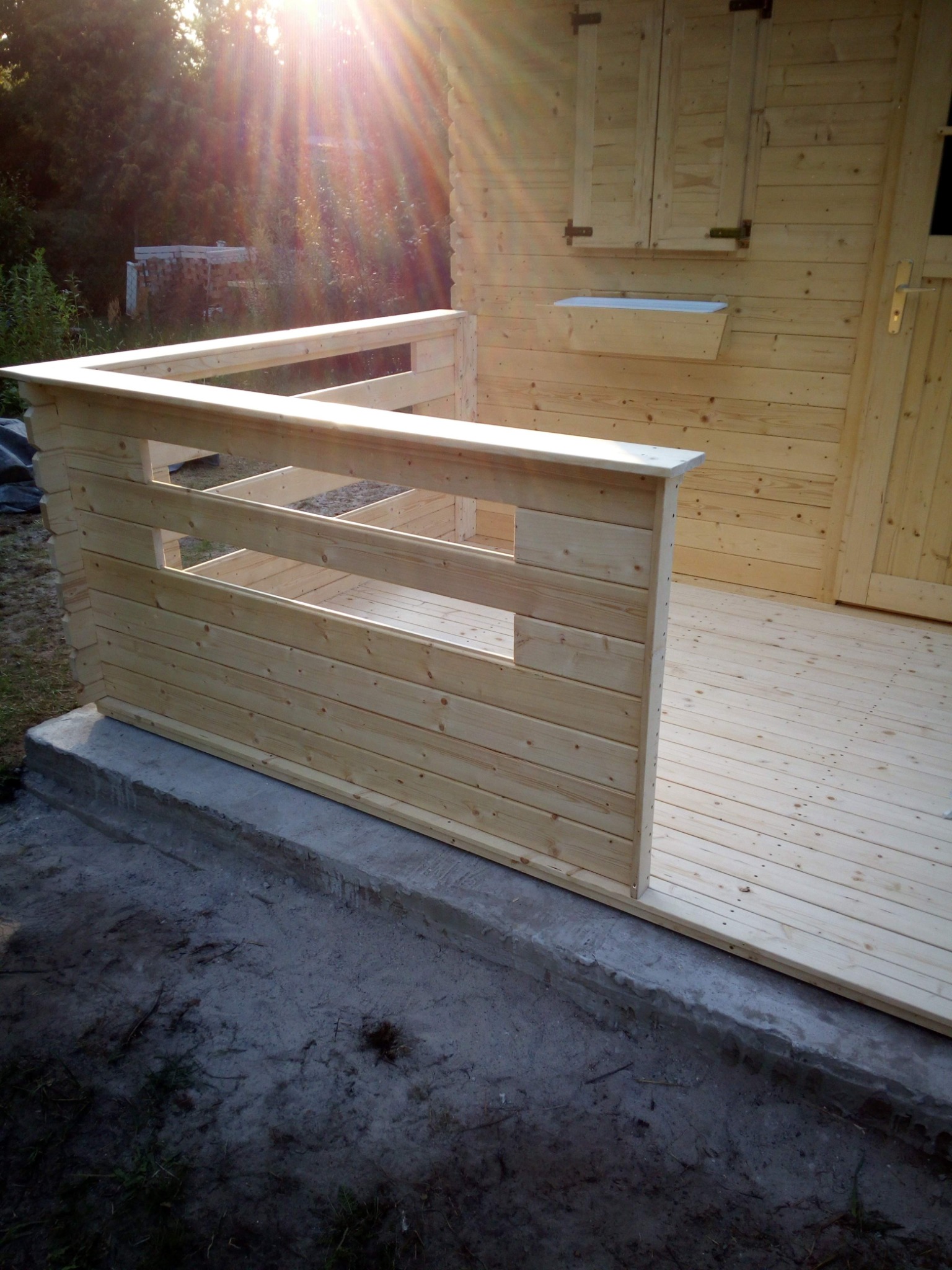 Nowo wybudowany, surowy taras z jasnego drewna z balustradą i podłogą, widoczny fragment elewacji domku z oknem i donicą, w tle zieleń.